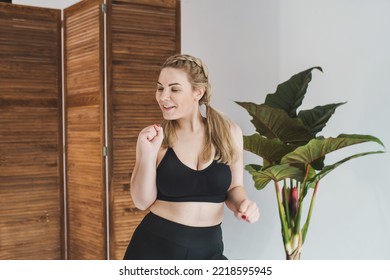 Happy Plus Size Woman In A Black Sports Top And Leggings Dancing. Chubby Blonde Is Engaged In Fitness. Curvy Girl Posing  In A Bright Room 