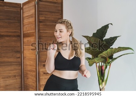 Happy plus size woman in a black sports top and leggings dancing. Chubby blonde is engaged in fitness. Curvy girl posing  in a bright room 
