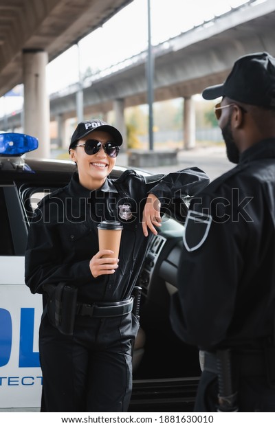 happy\
policewoman with paper cup looking at african american colleague\
while leaning on patrol car on blurred\
background