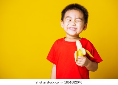 Happy Portrait Asian Child Or Kid Cute Little Boy Attractive Smile Wearing Red T-shirt Playing Holds Peeled Banana For Eating, Studio Shot Isolated On Yellow Background