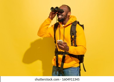 Happy. Portrait of a cheerful young african-american tourist guy with bag looking with binoculars isolated on yellow studio background. Preparing for traveling. Resort, human emotions, vacation.