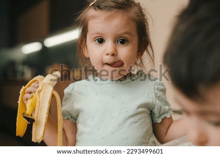 Happy portrait of a cute little baby girl holding peeled banana for eating, sticking out tongue, looking at camera. Healthy food. Happy family. Sweet food.