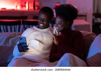 Happy Positive African Couple Relax On Sofa At Home With Smartphone, Use Phone To Video Chat With Family During Lockdown, Black Man And Woman Watch Funny Internet Video On Cellphone. Selective Focus