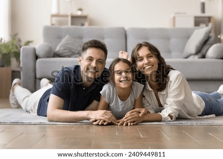 Happy positive attractive parents and cute tween girl kid resting together on heating floor at home, looking at camera, smiling. Caring mom, dad and daughter lying on bellies, posing for portrait