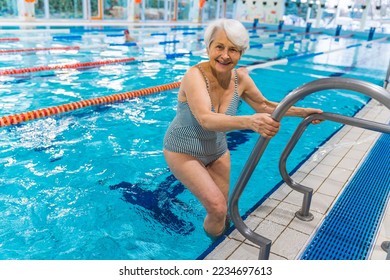 Happy positive caucasian senior adult woman in a striped swimming suit using metal stairs to get into the pool. Full-length indoor shot. Looking at camera. High quality photo - Powered by Shutterstock