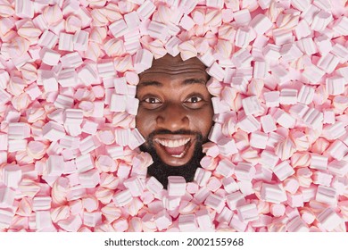 Happy positive dark skinned bearded man smiles broadly poses among delicious pink soft marshmallows Human head through tasty dessert background. Food background. Sweet dessert. Sugar addiction