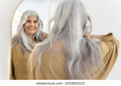 Happy positive elderly Hispanic woman mirror reflection and back view. Cheerful senior model touching long naturally grey hair, looking at herself, laughing, smiling