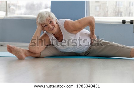 A happy, positive elderly woman does splits and yoga exercises in a bright sportive room. active old age concept
