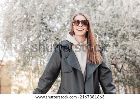 Happy positive fashion beautiful girl with stylish eyewear in a fashionable black leather jacket with a hoodie walking and enjoying near a blooming tree in the spring city. Female happiness mood