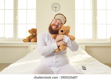 Happy Positive Funny Fat Grown Up Adult Man With Ginger Beard In White Pajamas And Sleep Mask Sitting On Bed In Bedroom, Cuddling Favorite Teddy Bear Like A Baby And Smiling. Immature Behavior Concept