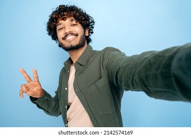 Happy positive Indian or Arabian excited guy with gladden face expression, in casual shirt, doing selfie shot on cellphone and smiles into the camera, shows peace symbol, isolated blue background