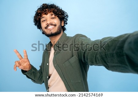 Happy positive Indian or Arabian excited guy with gladden face expression, in casual shirt, doing selfie shot on cellphone and smiles into the camera, shows peace symbol, isolated blue background