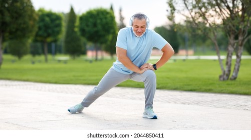 Happy Positive Mature Sportsman During Outdoor Workout, Senior Man Wearing Headphones And Sports Outfit Warming Up Muscles, Doing Side Squat On One Leg, Enjoying Active Lifestyle Outside In Park