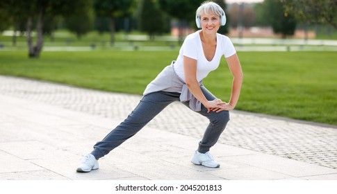 Happy Positive Mature Sportswoman During Outdoor Workout, Senior Woman Wearing Headphones And Sports Outfit Warming Up Muscles, Doing Side Squat On One Leg, Enjoying Active Lifestyle Outside In Park