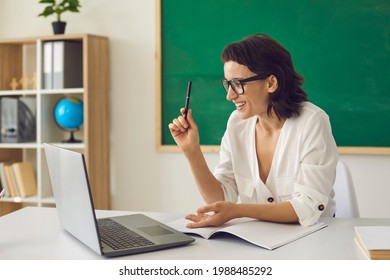 Happy Positive School Teacher Having Online Class. Smiley Young Woman In Glasses Sitting At Desk In Classroom And Using Laptop Computer To Teach Her Students Via Video Conference