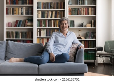 Happy Positive Senior Homeowner Lady Resting On Soft Comfortable Couch At Home, Looking At Camera, Smiling, Posing In Living Room Interior With Bookshelves Background