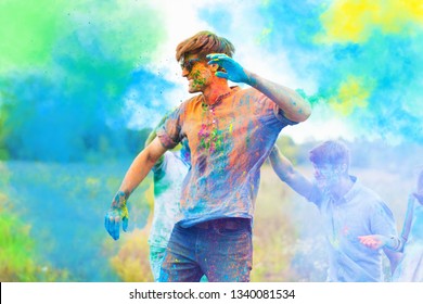 Happy positive smiling fun man wearing blue sunglasses all stained with colorful paint celebrating Holi festival party with friends. On color dust smoke powder cloud background in park field outdoor. 