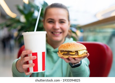 Happy positive smiling girl, young glad blurred woman is holding big juicy fat burger, hamburger and glass of soda in shopping mall on food court. Fast food restaurant, junk unhealthy meal concept.