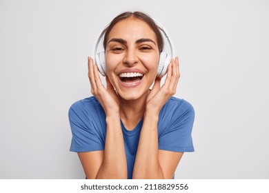 Happy positive woman with dark combed hair keeps hands on stereo headphones enjoys listening favorite music wears blue t shirt isolated over white background. People lifestyle hobby concept.