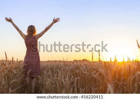 Happy positive woman full of vitality raising hands and facing the sun in a wheat field at sunrise