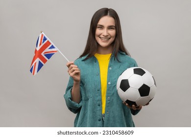 Happy positive woman holding in hands flag of United Kingdom and football ball, supporting favourite team on championship, wearing casual style jacket. Indoor studio shot isolated on gray background. - Powered by Shutterstock