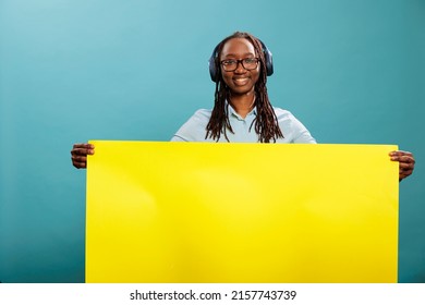 Happy positive woman holding yellow paper banner sign while having wireless modern headphones on blue background. Confident young adult person with electronic earphones and empty advertisement placard