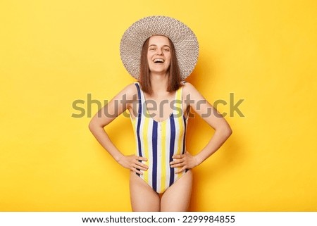 Happy positive woman wearing striped swimming suit and sun hat isolated on yellow background, enjoying her holiday, keeps hands on hips, looking at camera.
