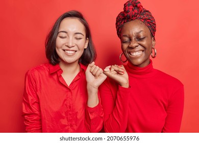 Happy positive women hook each others little fingers in conciliation or friendship smile toothily keep eyes closed stand closely against bright red background wear casual clothes. Relationship concept