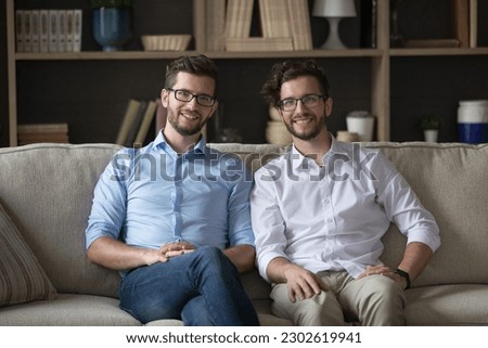 Happy positive young adult identical twin men in glasses sitting together on couch, looking at camera, smiling, meeting at home for family event, keeping good close relationships. Indoor portrait