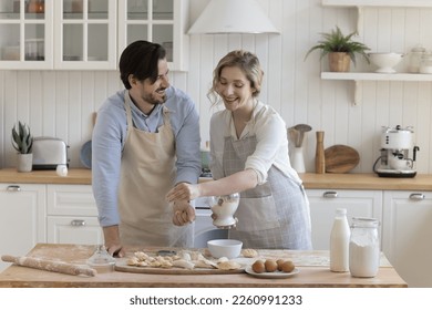 Happy positive young couple in love wearing aprons, enjoying bakery food preparation, baking pies in home kitchen, sifting flour on dough at table with ingredients, talking, smiling - Powered by Shutterstock