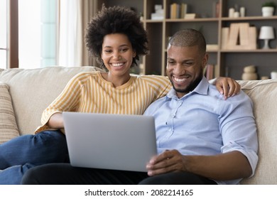 Happy positive young couple making video call, using laptop computer for online talk, looking at screen, smiling, laughing, resting on cozy sofa together at home. Communication concept