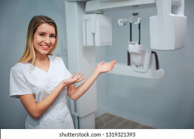 Happy positive young dentist stand in x-ray room and point on machine. Alone in white robe. - Shutterstock ID 1319321393