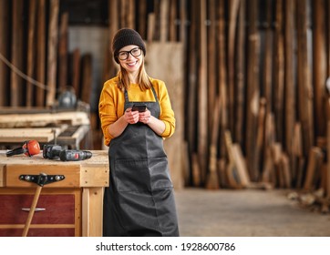 Happy positive young female woodworker in apron messaging on mobile phone and smiling at camera while standing near workbench in joinery workshop - Powered by Shutterstock