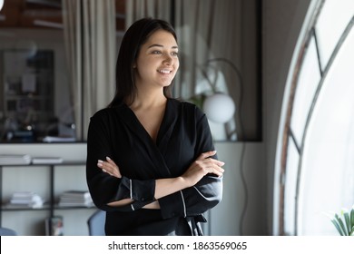 Happy Positive Young Indian Lady Standing By Window In Office Looking At Distance In Good Mood, Drawing Future Perspectives In Mind, Dreaming Of Career Growth, Imagining New Way Of Developing Business