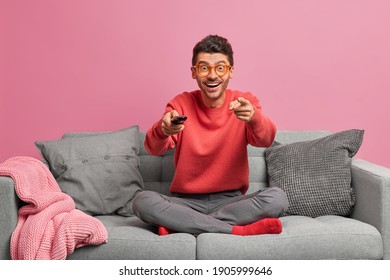 Happy positive young man watches something on TV holds remote control sits crossed legs on comfortable sofa in living room isolated over pink background. People lifestyle entretainment concept