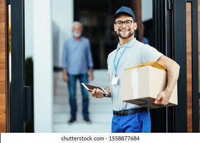 Happy Postal Worker With Digital Tablet Carrying Packages And Looking At Camera While Making Home Delivery.  