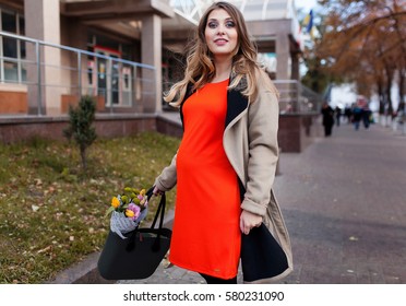 Happy pregnancy concept. Young beautiful fashion woman walking on the street sunny spring day with flowers on her bag.