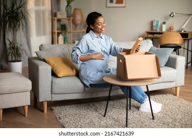 Happy Pregnant African American Female Unpacking Cardboard Moving Box Holding Photo Phrame Sitting On Sofa At Home. Pregnancy Lifestyle, Shopping And Delivery Service
