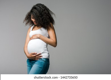happy pregnant african american girl touching belly while standing on grey 