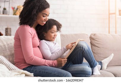 Happy Pregnant Afro Mom And Her Sweet Little Daughter Spending Time Together At Home, Reading Book, Choosing Name For Future Baby