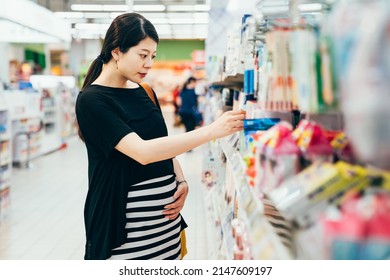 Happy pregnant asian chinese woman looking at product at grocery store. side view smiling future mom shopping in supermarket and reading product information on shelf. Customer buying in hypermarket