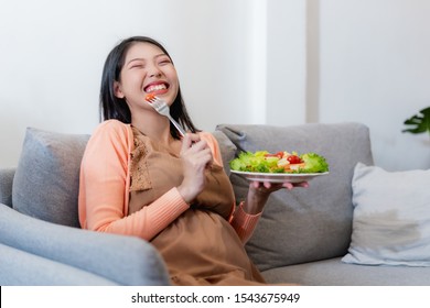 Happy Pregnant Asian Woman Sitting And Eating Natural Vegetable Salad Healthy Food And Sitting On Sofa