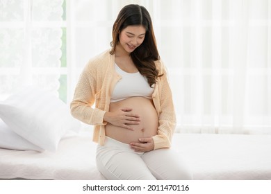 Happy Pregnant Asian Woman sitting on bed holding and stroking her big belly at home,Pregnancy of young woman enjoying with future life relax at home,Motherhood and Pregnant Concept,Soft focus - Powered by Shutterstock