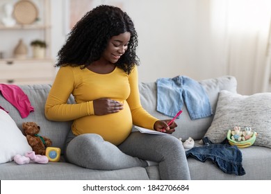 Happy Pregnant Black Woman Sitting On Sofa With Notebook And Pen, Making Shopping List Before Delivery, Free Space. Pretty African American Single Mother Getting Ready For Baby Coming, Home Interior