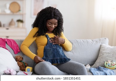 Happy Pregnant Black Woman Sitting On Sofa With Notebook And Pen, Looking At Small Dress For Baby, Free Space. Pretty African American Single Mother Getting Ready For Baby Coming, Home Interior