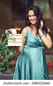 A Happy Pregnant Brunette In A Green Dress Is Holding A Birthday Cake With A 30th Birthday Candle. Pregnant Woman Expecting A Baby And Celebrating Her Birthday.