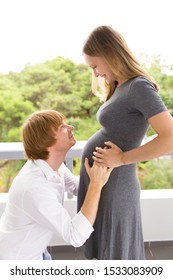 Happy Pregnant Couple Enjoying Expecting Baby. Side View Of Joyful Man And Woman Holding Baby Bump. Looking At Each Other, Smiling. Enjoying Pregnancy Concept