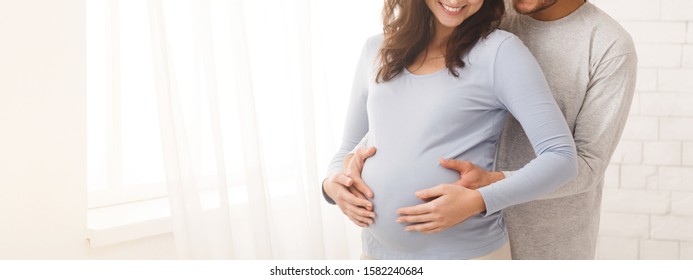 Happy pregnant couple hugging in front of the window at home, panorama with free space