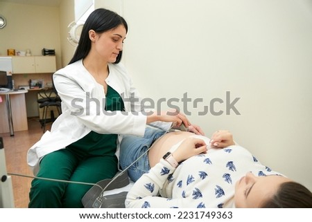 Happy pregnant female visiting her own gynecologist in medical clinic. Doctor examining the baby with stethoscope and ctg graph. Fetal heart monitoring. Cardiotocography.               