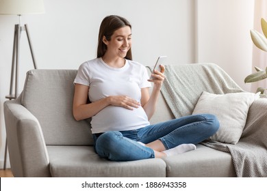 Happy pregnant millennial woman using smartphone at home, browsing pregnancy app while sitting on couch in living room, smiling expectant lady looking at phone screen and touching belly, free space - Powered by Shutterstock